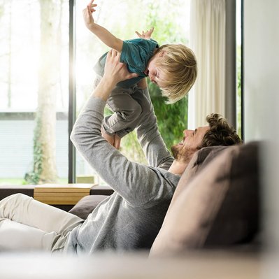 Mann hebt lachendes Kind hoch, helle Wohnzimmerumgebung. Darstellung von Wärme und Freude durch effizientes Heizen mit Wärmepumpe.