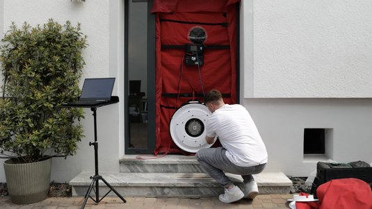 Demonstration des Blower-Door-Tests. Mann justiert Ventilator an der Abdichtung, Laptop zur Kontrolle der Messdaten, um Luftdichtheit zu testen.