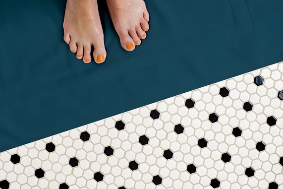 Bild zeigt auf der einen Hälfte Füße auf einem glatten Boden. Auf der anderen Hälfte grenzt eine Mosaik-Fliesenboden an. Es geht um den Stromverbrauch der Fußbodenheizung.