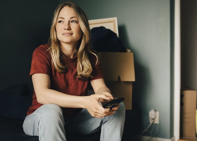 Eine Frau mit Smartphone sitzt zwischen Kartons in der Wohnung. Sie wartet beim Umzug auf die Strom Endabrechnung und finalen Zählerstand.